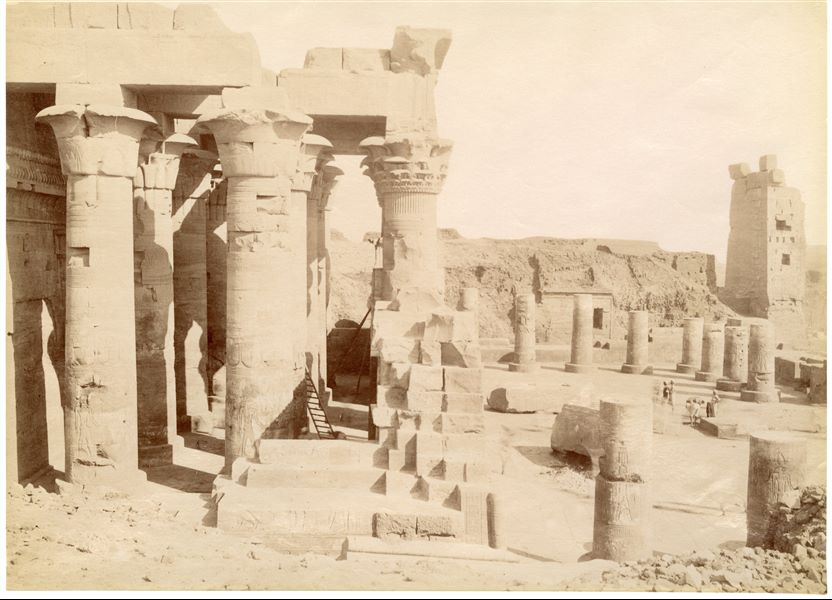 The photograph shows the external hypostyle hall (built by Ptolemy XII) and the front courtyard (built by Augustus) from the Temple of Sobek and Haroeris at Kom Ombo seen from the north-west. In the background, on the right, the entrance pylon to the sacred precinct is visible. Based on the calligraphic style of the caption, the shot can be attributed to Antonio Beato. 