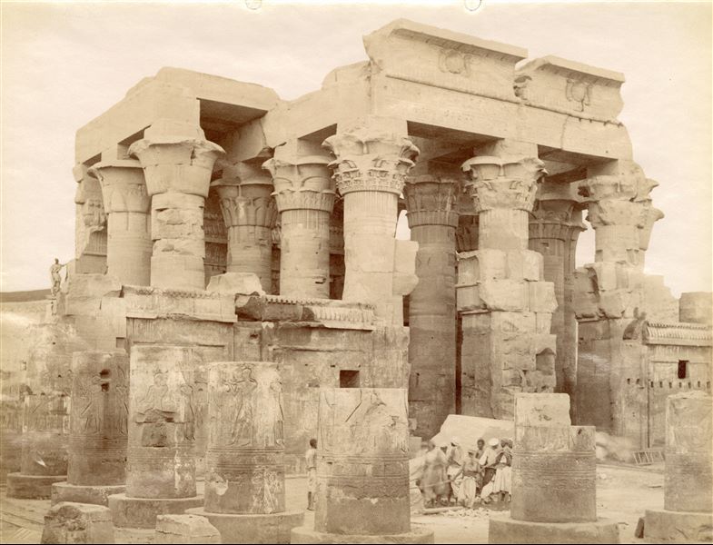 The photograph depicts the external columned hall (built by Ptolemy XII) of the Temple of Sobek and Haroeris at Kom Ombo, seen from the courtyard in front of it (that of Augustus). A group of men are pulling something that is hidden by a column, and an egyptian has climbed to the top of the west (left) wall. Based on the calligraphic style of the caption, the image can be attributed to Antonio Beato.