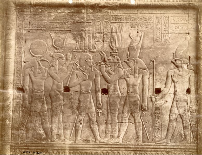 The photograph shows a sacred scene from the interior walls of the Temple of Sobek and Haroeris at Kom Ombo, a bas-relief located in the area consecrated for Horus. Ptolemy XII receives the Ankh and crown from the gods Thoth, Bastet, Isis in the presence of Horus and Ra. 