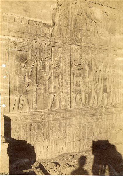 The photograph shows wall scenes from the Temple of Sobek and Haroeris at Kom Ombo, where Pharaoh Ptolemy VIII is in the presence of some deities. The shadow of the photographer and camera can be seen at the bottom. 