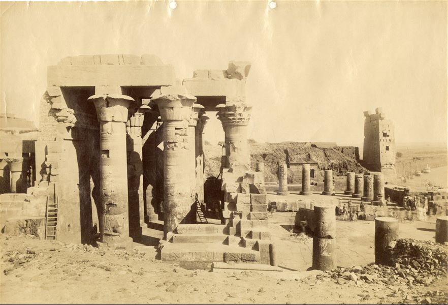 Northern view of the front courtyard and hypostyle hall from the remains of the Temple of Sobek and Haroeris at Kom Ombo. The image (which had a long exposure) shows several people moving on the right, presumably to clear the temple of debris, and has resulted blurred and out of focus. In the background on the right, the Nile and some feluccas, the typical Egyptian boats used to sail along the river.  
