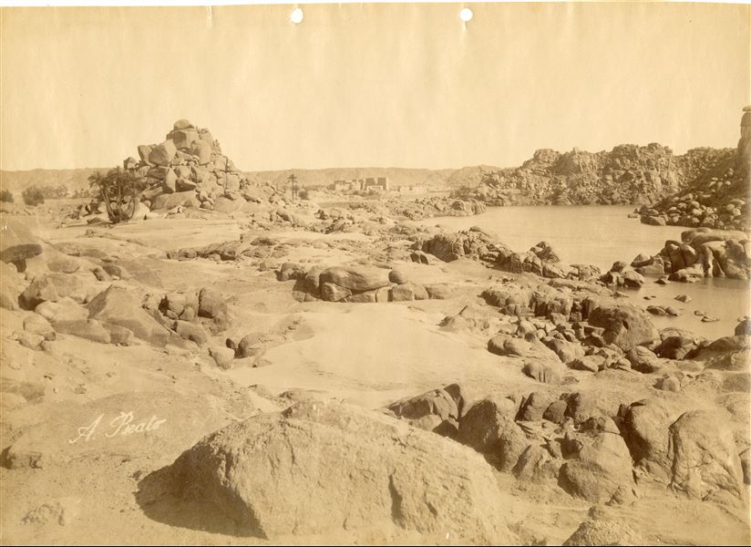 The photograph shows a view of the landscape and rocks emerging from the Nile at the First Cataract, near Aswan. In the background, the Temple Complex of Isis on the island of Philae can be seen. Today, the First Cataract has been submerged by Lake Nasser, created as a result of the construction of the Aswan Dam in the 1960s, and the Temple Complex was moved to a neighbouring island because it was threatened by the same lake. The author's signature is visible at the bottom left. 
