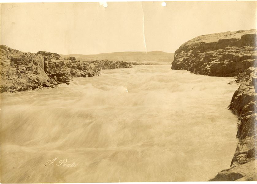 Panoramica del paesaggio desertico di Assuan, interrotto dal corso impetuoso del Nilo all'altezza della Prima Cateratta. In quest’area venne negli anni ‘60 costruita la diga di Assuan. In basso a sinistra la firma dell’autore.