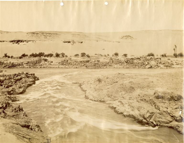 The photograph shows the rapids of the First Cataract of the Nile at Aswan. The author's signature (slightly faded) is written in the lower left corner. 