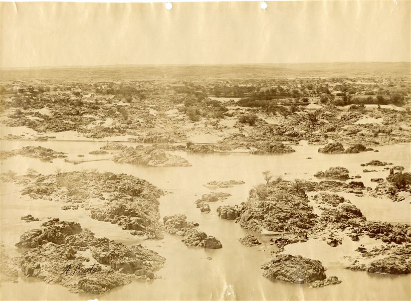 The photograph shows a view of the panorama and the rocks emerging from the Nile at the First Cataract, near Aswan. Today, the First Cataract has been submerged by Lake Nasser, which was created as a result of the construction of the Aswan Dam in the 1960s. The author's signature is visible at the bottom left. 