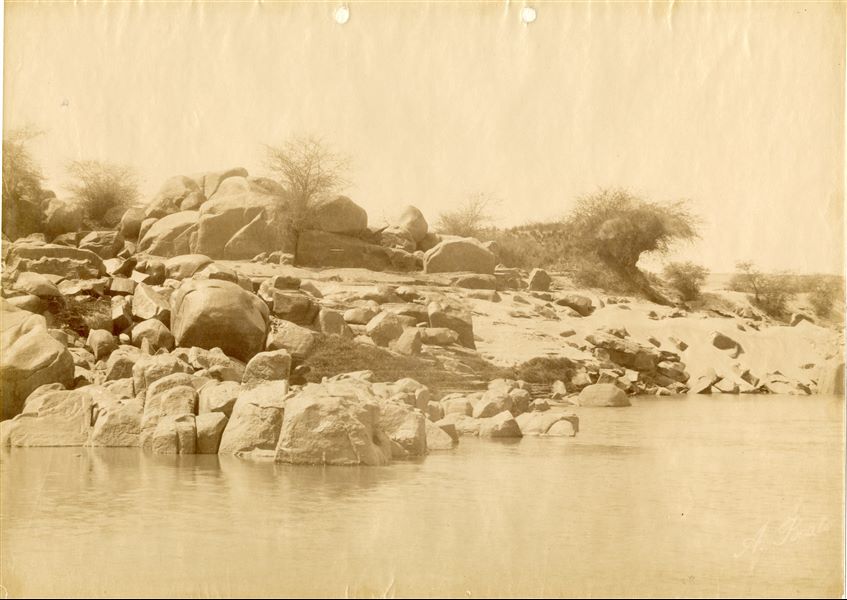 The photograph illustrates the banks of Chelal (caption on the back of the image), at the height of the First Cataract of the Nile, near Aswan. The author's signature (slightly faded) is written at the bottom right.. 