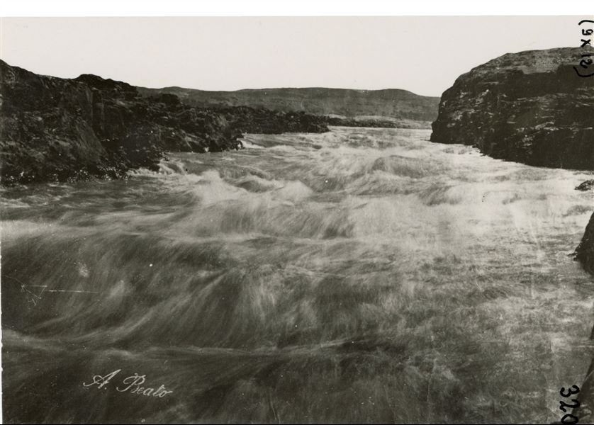 The photographed landscape shows the rapids of the First Cataract of the Nile near Aswan, and the rocky landscape of Upper Egypt. The author's signature is clearly visible at the bottom left.