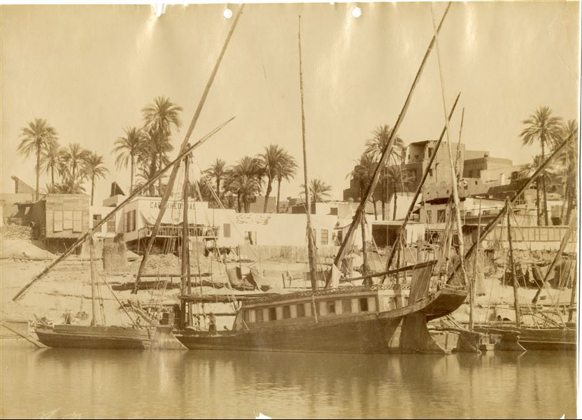 The photograph depicts a view of the town of Aswan on the banks of the Nile, showing some boats and houses. The author's signature can be found at the bottom left. 