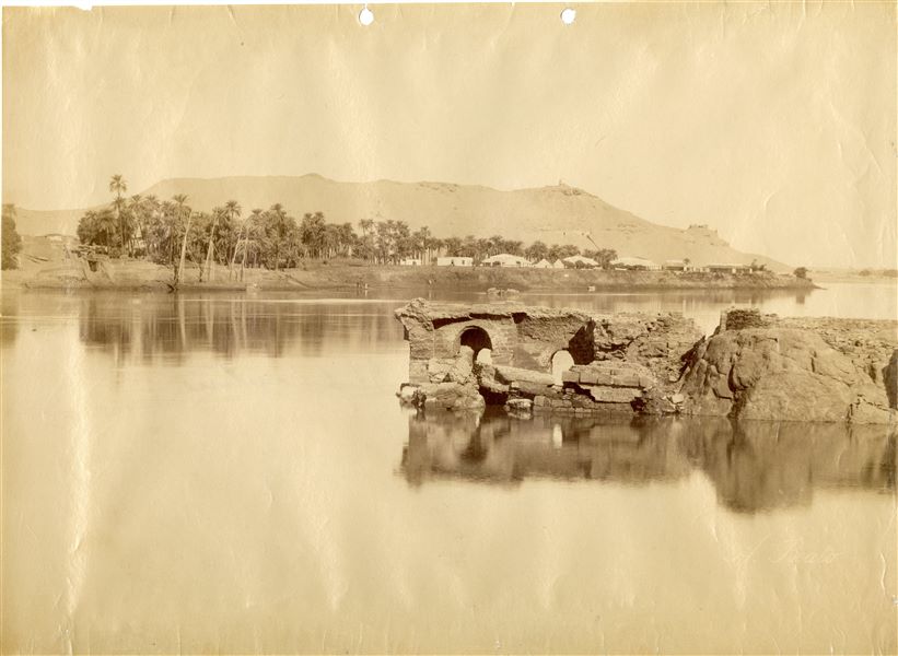 The image captures a view of the Elephantine Island on the Nile. The mountains of Aswan are in the background, with the southern slope of Qubbet el-Hawa, where the tomb of Sheikh Ali Abu el-Hawa is visible on its summit. The author's signature can be found at the bottom right. 