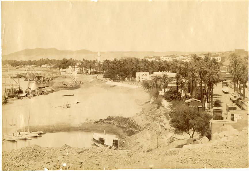 The photograph shows a panorama of the city of Aswan along the banks of the Nile. With some moored boats and the riverbank (left), as well as a minaret (centre) and a railway (right).  