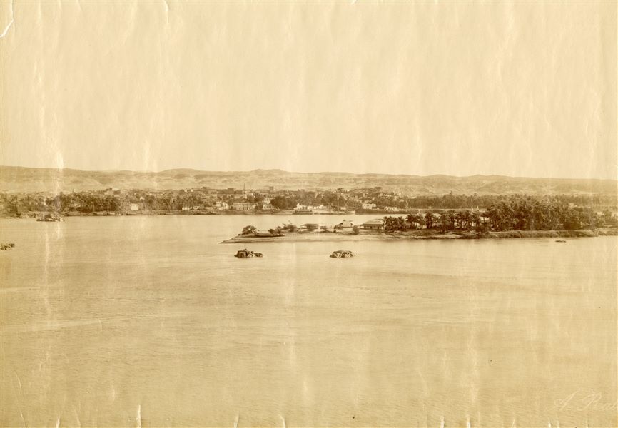 Panorama showing a small town on the banks of the Nile, probably Aswan. In the centre, a small island. Photograph taken from the west bank of the river. The author's signature (faded) is at the bottom right. 