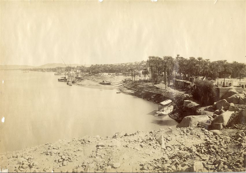 The photograph depicts a view of the Nile in a northerly direction, photographed in the vicinity of Aswan and the First Cataract. On the right bank, a railway convoy on rails and a station, with people waiting, visible through the palm trees. The author's signature (faded and barely legible) is at the bottom right.