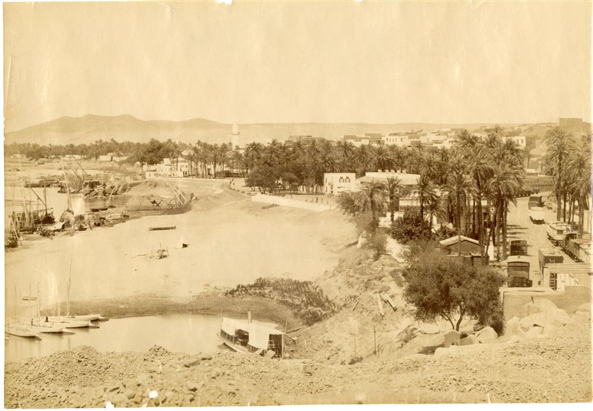 The photograph shows a panorama of the city of Aswan along the banks of the Nile. With some moored boats and the riverbank (left), as well as a minaret (centre) and a railway (right).  