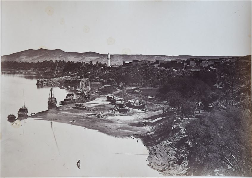 The photograph shows a view of the town of Aswan on a palm-fringed bank of the Nile where some boats are moored. Other boats are pulled ashore, presumably for some carpentry work. The author's signature is visible at the bottom centre. 