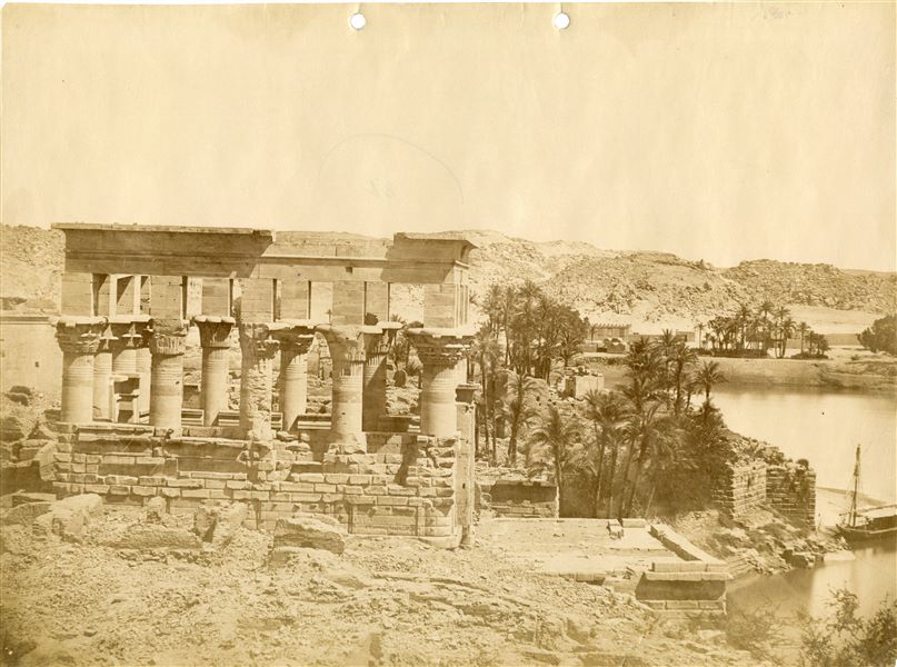 Photograph of Trajan's Kiosk in Philae in its original location, and the eastern shore of the island, with buildings, palm groves and a moored boat. 