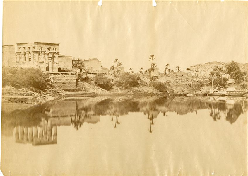 Photograph representing a side view of the temple complex of Isis at Philae. The Kiosk erected by Emperor Trajan in the 2nd century AD stands out on the left.  In the foreground, the Nile with two feluccas moored near the small island. The author's signature is on the left. 