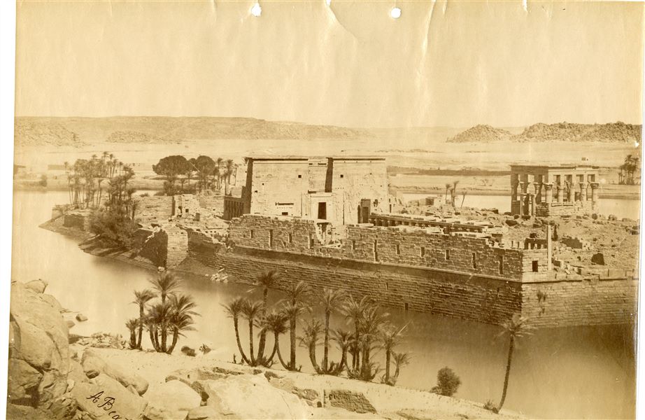 The photograph shows a view of the island of Philae from the southwest, where the façade of the Temple of Isis and Trajan’s Kiosk, together with the large retaining structures and the west portico stand out. In the foreground, the shoreline of the island of Bigeh and the Nile, with the Upper Egyptian landscape visible in the background. The temple complex was relocated in the 1970s to the nearby Agilkia island, following the construction of the Aswan Dam and the formation of Lake Nasser, which would have submerged it. The author’s signature is at the bottom left.