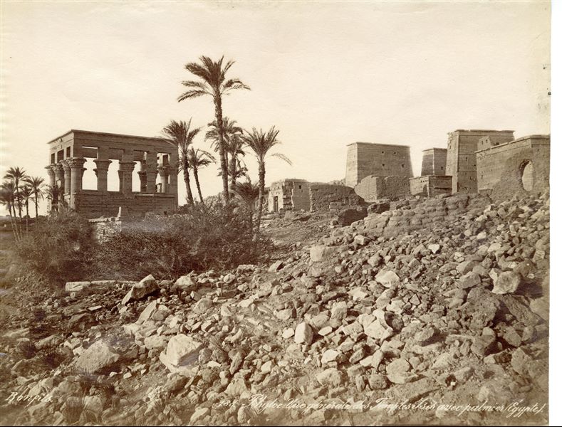 View of the northern ruins of the temple complex of Isis at Philae, with mud-brick structures (no longer surviving), together with Trajan’s Kiosk in its original position, and some palm trees. The author's signature is visible at the bottom left. 