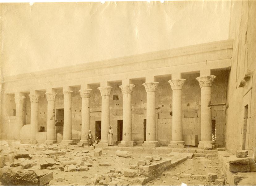 The photograph shows the colonnade of the Temple of Isis in Philae. In the centre, three egyptians pose for the photographer.  Part of the entrance pylon can be seen on the right. The author's signature is at the bottom left. 