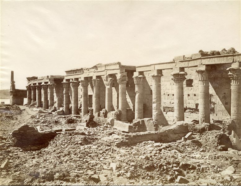 View of the western colonnade from the temple complex of Isis at Philae, in its original position, with the Kiosk of Pharaoh Nectanebo and the Nile in the background. The temple was relocated in the 1970s to the nearby  Agilkia island following the construction of the Aswan Dam and the formation of Lake Nasser, which would have submerged it. The author's signature is visible at the bottom right. 
