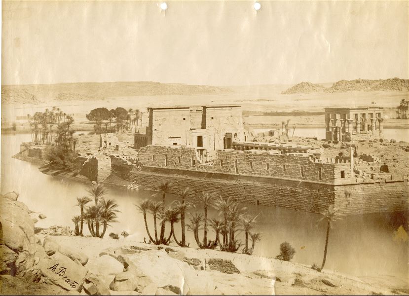 The photograph shows a view of the island of Philae from the southwest, where the façade of the Temple of Isis and Trajan’s Kiosk, together with the large retaining structures and the west dromos stand out. In the foreground, the shoreline of the island of Bigeh and the Nile, with the Upper Egyptian landscape visible in the background. The temple complex was relocated in the 1970s to the nearby Agilkia island, following the construction of the Aswan Dam and the formation of Lake Nasser, which would have submerged it. The author’s signature is at the bottom left. 