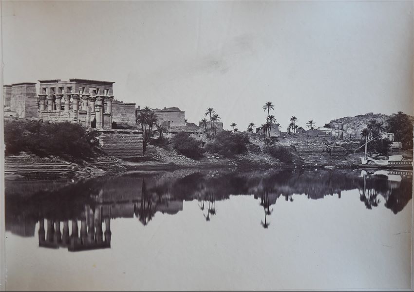 Fotografia del complesso templare di Philae visto dal Nilo. A sinistra, è ben visibile il Chiosco di Traiano. A destra invece è presente una feluca ormeggiata. La firma dell’autore è posta a sinistra, nel riflesso sull’acqua.  