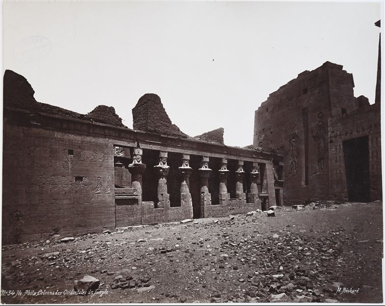 View of the western part of the second courtyard from the Temple of Isis on the island of Philae. The second pylon is visible on the right, while on the left is the Mammisi, with the portico displaying columns with Hathoric capitals. The author's signature is at the bottom right.  