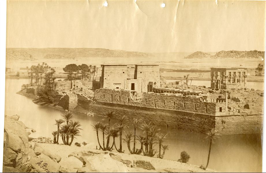 The photograph shows a view of the island of Philae from the southwest, where the façade of the Temple of Isis and Trajan’s Kiosk, together with the large retaining structures and the west portico stand out. In the foreground, the shoreline of the island of Bigeh and the Nile, with the Upper Egyptian landscape visible in the background. The temple complex was relocated in the 1970s to the nearby Agilkia island, following the construction of the Aswan Dam and the formation of Lake Nasser, which would have submerged it. The author’s signature is at the bottom left.