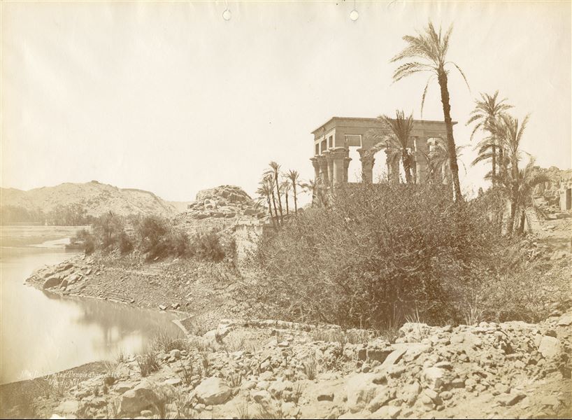 The photograph depicts the Kiosk erected by Emperor Trajan, seen from the north, rising among the vegetation of the banks of the Nile. In the background on the right, the ruins of the temple complex dedicated to the goddess Isis on the island of Philae. The author's signature is visible at the bottom right. 
