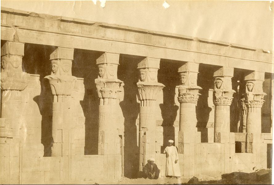 The photograph shows the entrance to the Mammisi of Horus (a temple dedicated to the birth of Horus, whom Isis gave birth to) located near the second pylon in the temple complex of Isis at Philae, with two egyptians staring at the camera lens. The ground still partially covers part of the elevated structure. The author's signature is visible at the bottom right. 