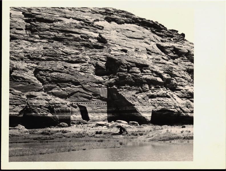 Photograph of the rock-cut chapel of Ellesiya in its original location and of the plain in front of the rock-cut chapel, in Nubia. Photographed shortly before the Nile waters (visible in the foreground) would begin to rise due to the construction of the Aswan Dam, which would flood the area. Photo taken in the mid-1960s, shortly before the rock-cut chapel was moved.