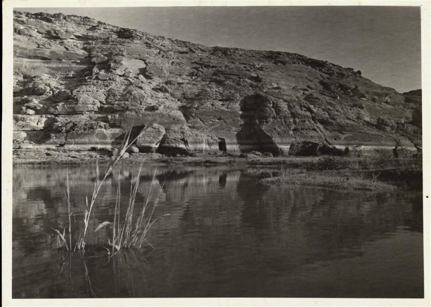 Photograph of the rock-cut rock-cut chapel of Ellesiya in its original location within the Nubian landscape. In addition to the mountain (in which the rock-cut chapel was carved), the Nile River can be seen in the foreground, which shortly afterwards, would begin to rise until it submerged the rock-cut chapel, due to the construction of the Aswan Dam. 