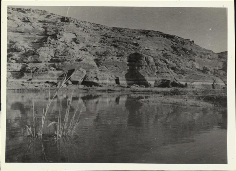 Photograph of the rock-cut rock-cut chapel of Ellesiya in its original location within the Nubian landscape. In addition to the mountain (in which the rock-cut chapel was carved), the Nile River can be seen in the foreground, which shortly afterwards, would begin to rise until it submerged the rock-cut chapel, due to the construction of the Aswan Dam. 

