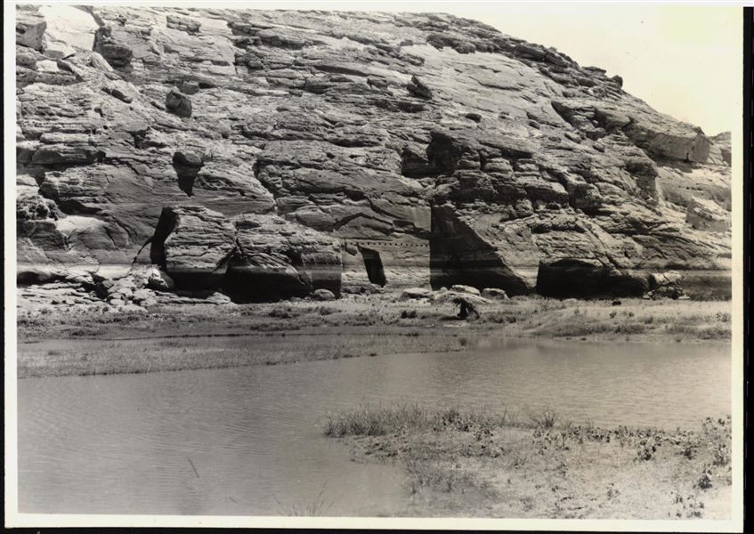 Photograph of the rock-cut rock-cut chapel of Ellesiya in its original location within the Nubian landscape. In addition to the mountain (in which the rock-cut chapel was carved), the Nile River can be seen, which shortly afterwards, would begin to rise until it submerged the rock-cut chapel, due to the construction of the Aswan Dam.