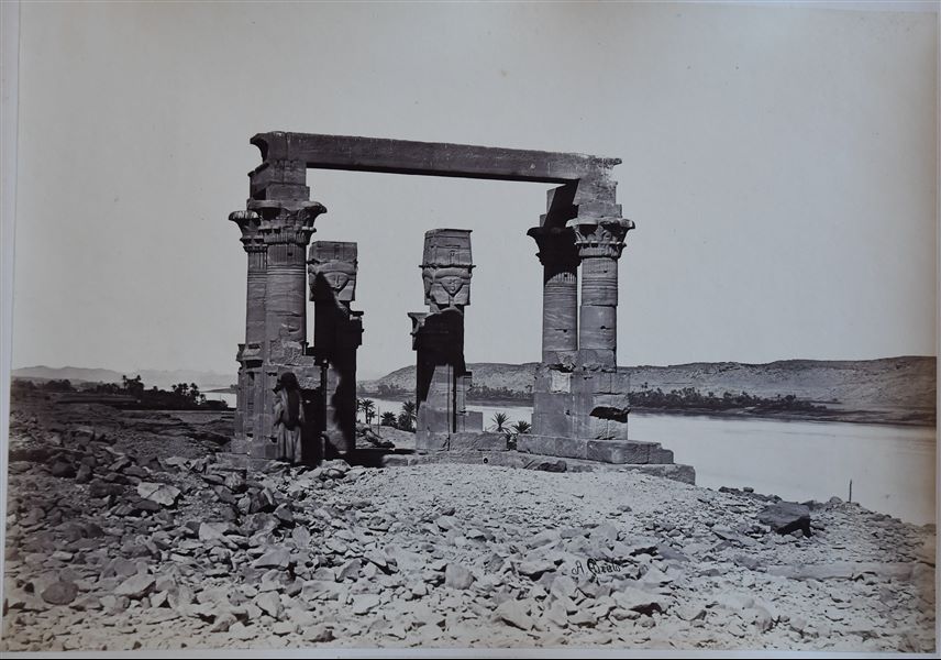 View of the remains of the Roman Period (3rd century A.D.) kiosk in the Temple of Isis at Qertassi in Nubia, on the banks of the Nile. Only a few columns remain, two with Hathor capitals, four with papyrus capitals. A young Nubian is posing for the camera in front of a column. The author's signature is at the bottom right. 