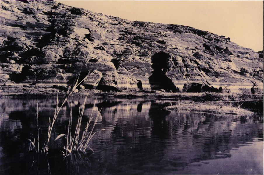 Photograph of the rock-cut Temple of Ellesiya in its original location within the Nubian landscape. In addition to the mountain (in which the temple was carved), the Nile River can be seen in the foreground, which shortly afterwards, would begin to rise until it submerged the temple, due to the construction of the Aswan Dam.
