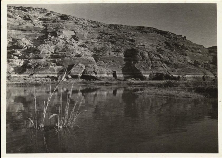 Photograph of the rock-cut Temple of Ellesiya in its original location within the Nubian landscape. In addition to the mountain (in which the temple was carved), the Nile River can be seen in the foreground, which shortly afterwards, would begin to rise until it submerged the temple, due to the construction of the Aswan Dam. 