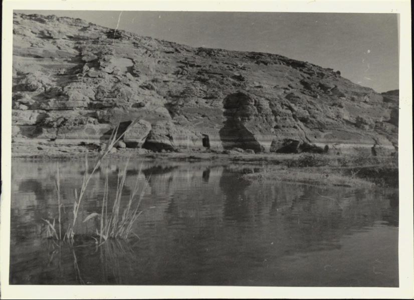 Photograph of the rock-cut Temple of Ellesiya in its original location within the Nubian landscape. In addition to the mountain (in which the temple was carved), the Nile River can be seen in the foreground, which shortly afterwards, would begin to rise until it submerged the temple, due to the construction of the Aswan Dam. 

