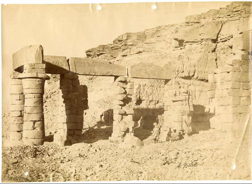 The photograph shows the ruins of the south side of the colonnaded hall of the Temple of Gerf Hussein, built by Ramesses II for the god Ptah. Three local inhabitants pose for the camera. In the 1960s (1962-1963), the remains of the colonnade were dismantled and moved to the area of New Kalabsha, a  location on higher  ground not far from Aswan. The part excavated into the rock however, was submerged by Lake Nasser. The author's signature is at the bottom right. 