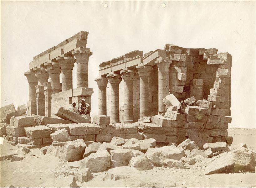 The photograph shows the ruins of the Roman temple of Maharraqa (seen from the eastern side) dedicated to the deities Isis and Serapis, in its original position, with some Nubians sitting on the blocks. The temple was moved following the construction of the Aswan Dam and the resulting formation of Lake Nasser, which would submerge it. It is now located at the new Wadi es-Sebua, where it was rebuilt in 1966-68. The author's signature is at the bottom right.