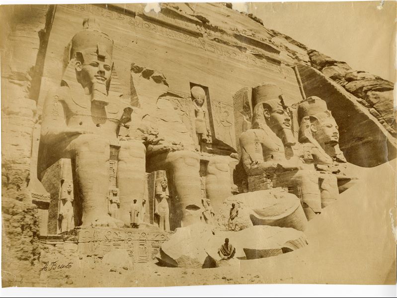 Veduta del monumentale ingresso del tempio maggiore di Abu Simbel, costruito dal faraone Ramesse II. Si nota ancora, sulla destra, la presenza di un cumulo di sabbia, che parzialmente copre il quarto colosso scolpito nella roccia. In posa, alcuni abitanti della zona. Negli anni ‘60 il tempio divenne il simbolo del salvataggio dei templi nubiani, minacciati dall’innalzamento delle acque del Lago Nasser in seguito alla costruzione della diga di Assuan. I due templi di Abu Simbel vennero infatti tagliati in blocchi e riassemblati su un’altura poco distante. La firma dell’autore è in basso a sinistra.