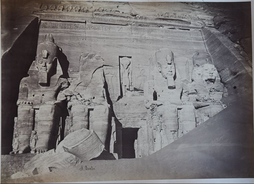 The photograph shows the exterior of the Great Temple of Abu Simbel, where one of the four colossal statues, together with the collapsed head of a second statue, are still partially covered by sand. The author's signature is clearly visible at the bottom.