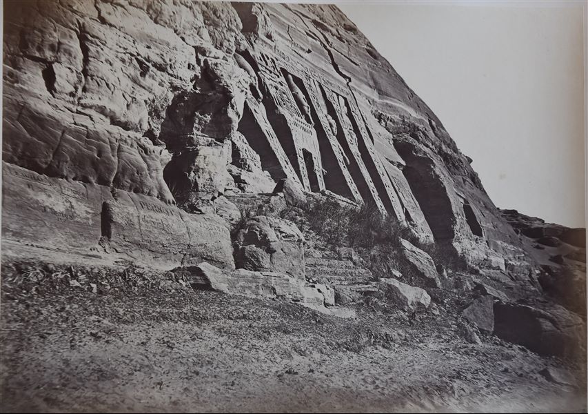 The photograph shows a view from below of the Small Temple of Abu Simbel. The colossal statues on the façade, portraying Ramesses II and his wife Nefertari can be seen. The rocky walls next to the temple are engraved with graffiti. The author's signature is at the bottom. 
