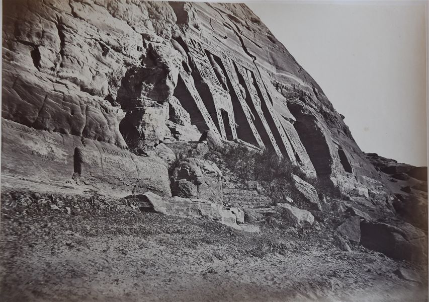 The photograph shows a view from below of the Small Temple of Abu Simbel. The colossal statues on the façade, portraying Ramesses II and his wife Nefertari can be seen. The rocky walls next to the temple are engraved with graffiti. The author's signature is at the bottom. 