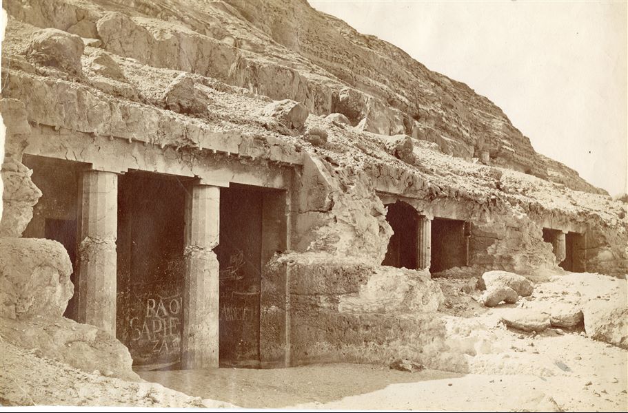 The photograph shows a series of tombs excavated in the mountain near Beni Hassan, a location in the 16th nome of Upper Egypt.  In the foreground, with modern graffiti is tomb No. 3 of Governor Khnumhotep II, and on the right, tomb No. 4 of Governor Khnumhotep IV. In the background, tomb No. 5. The author's signature is at the bottom right. 