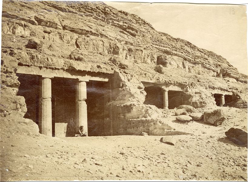 The photograph (like the previous one) shows a series of tombs excavated in the mountain near Beni Hassan, a location in the 16th nome of Upper Egypt.  In the foreground, with modern graffiti is tomb No. 3 of Khnumhotep II, and to the right, tomb No. 4 of Khnumhotep IV. In the background, Tomb No. 5. Photograph taken a few metres away from the point of the previous photograph. The author's signature is at the bottom right. 