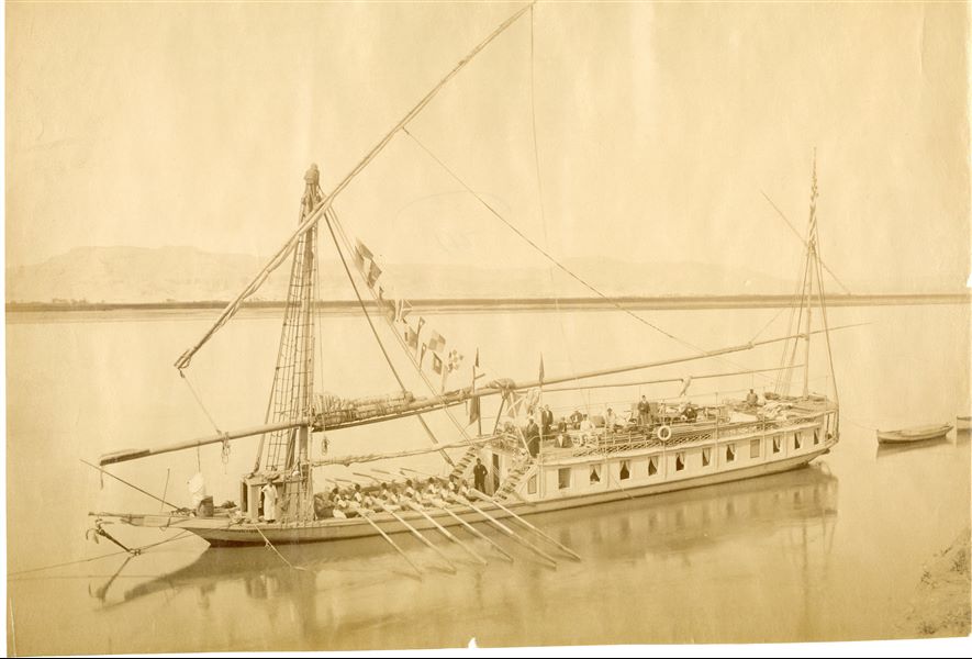 The photograph depicts a view of an entire cruise ship, with a group of tourists settled on the upper deck and the boatmen further down in the act of manoeuvring the boat, presumably ready to set sail or to dock. 