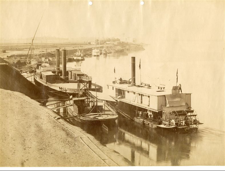 The photograph shows a number of steam-powered and sail boats moored on the banks of the Nile. 