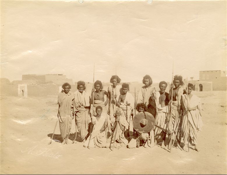 Nubian people posing for a photo, presumably warriors or hunters. Some are holding spears and swords, and a younger man holds a shield. The author’s signature, which is barely visible, is in the bottom left-hand corner. 