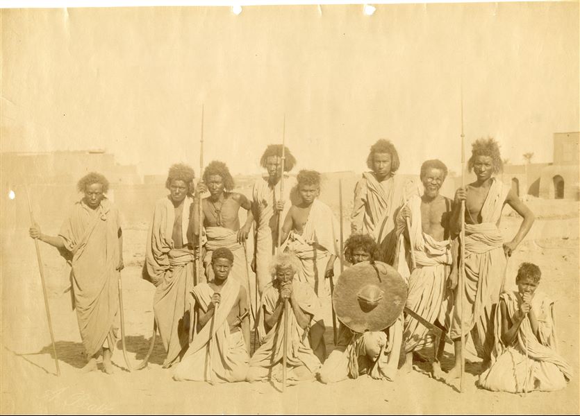 Popolazione nubiana in posa per uno scatto fotografico, presumibilmente dei guerrieri o cacciatori: alcuni tengono in mano lance e spade, un giovane mostra invece uno scudo. In basso a sinistra la firma poco visibile dell’autore. 